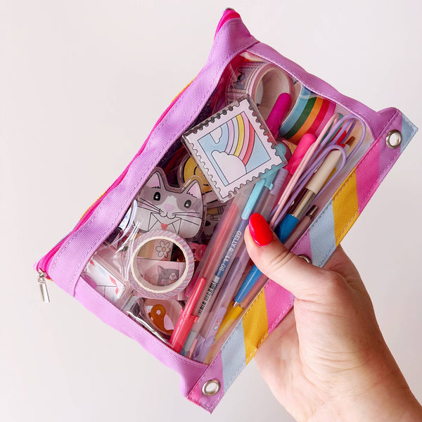 A hand with red nail polish holds a pink zippered pencil pouch filled with stationery items, shown against a plain light background.