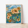 A framed art print featuring an expressive, textured painting of sunflowers in shades of yellow, orange, and brown against a vibrant blue and green background, leaning against a white wall on a light wood herringbone floor.