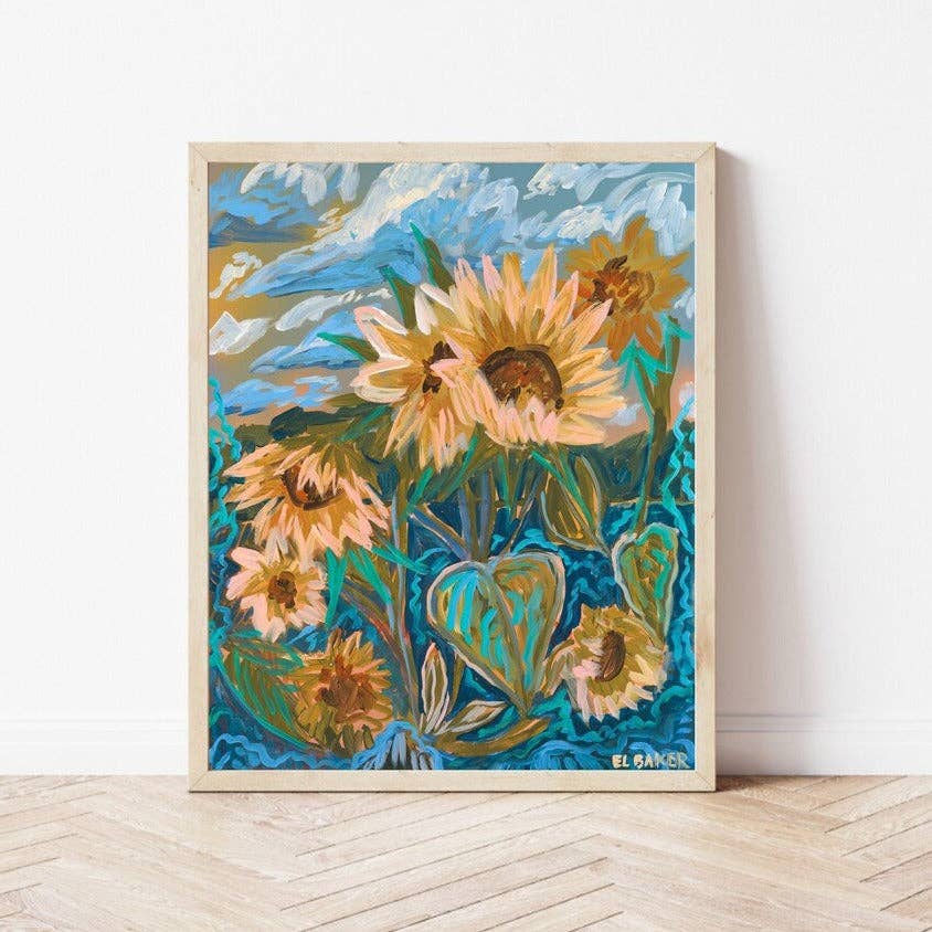A framed art print featuring an expressive, textured painting of sunflowers in shades of yellow, orange, and brown against a vibrant blue and green background, leaning against a white wall on a light wood herringbone floor.