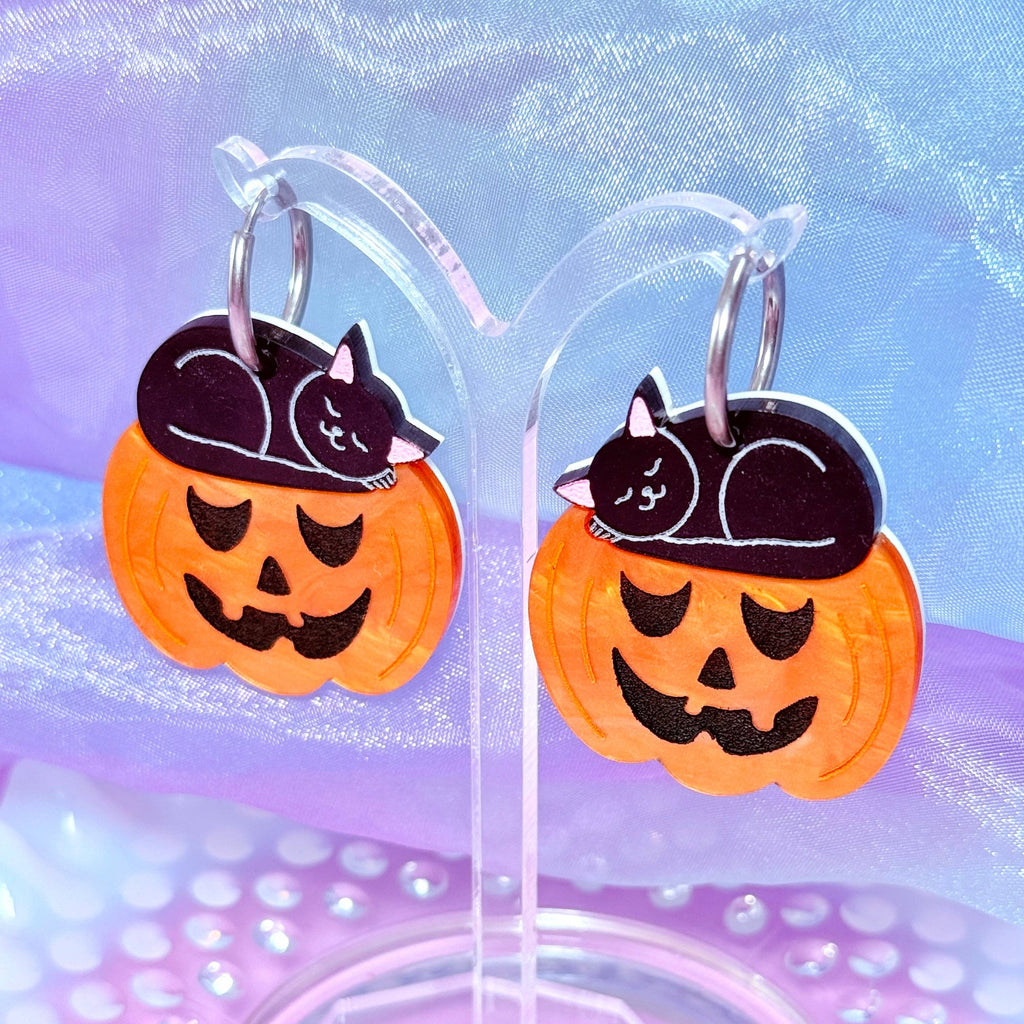 A pair of Halloween-themed dangle earrings displayed on a clear acrylic stand. Each earring features a bright orange pumpkin face with a black cat curled up sleeping on top, set against a purple and blue glittery background.