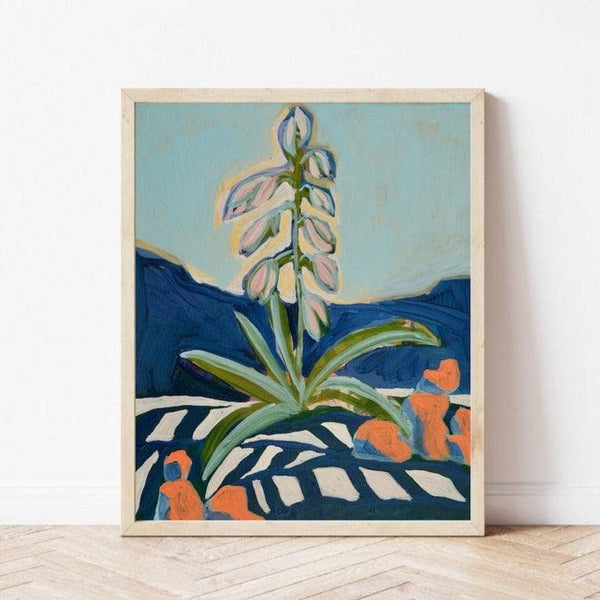 A framed art print featuring an abstract landscape painting with a central white flower, blue mountains, and orange accents, leaning against a white wall on a light wood floor.