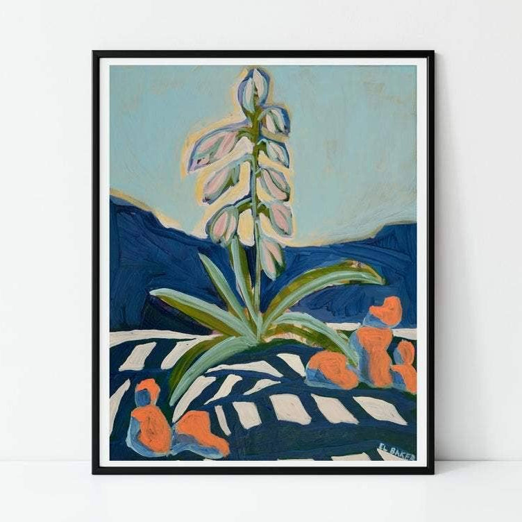 A framed art print featuring an abstract painting of a green plant with pale flowers against a light blue sky and dark blue hills, leaning against a white wall.