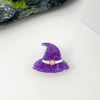 A small purple glitter witch hat accessory with a white band and a tiny pink center detail, placed on a white surface with a mossy rock and a white box in the background.