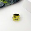 A small ring-shaped accessory with a Frankenstein-style face design, featuring a glittery yellow-green body, black hair, and gray side supports, photographed on a white surface with decorative rocks and a white box in the background.