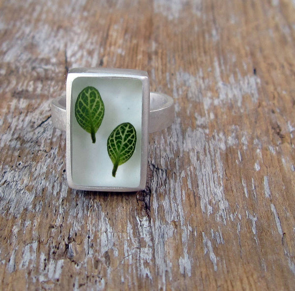 A sterling silver ring with a polished resin top featuring two green leaves, set on a wooden surface.