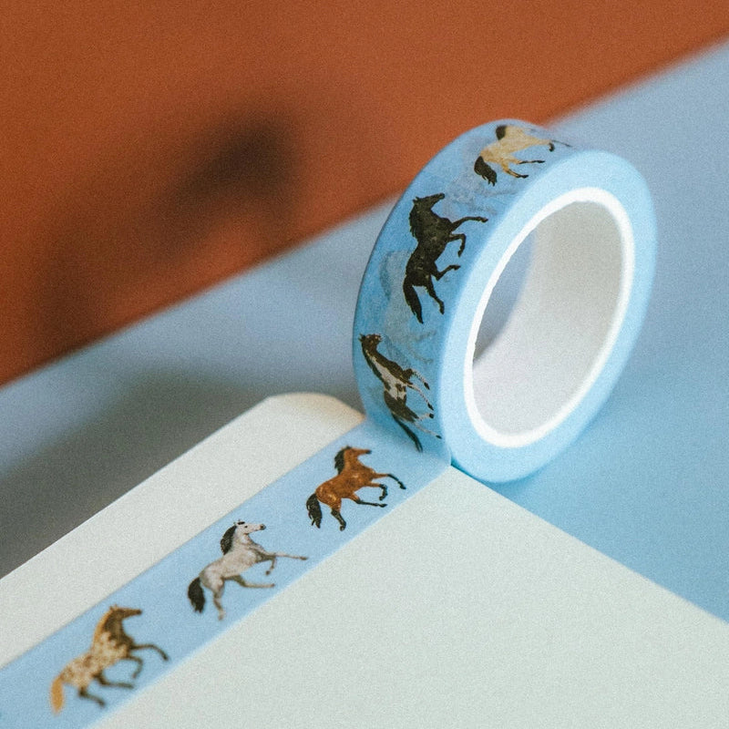 A roll of light blue washi tape with a repeating pattern of running horses in black, brown, and white, resting on a light blue surface next to a notebook.