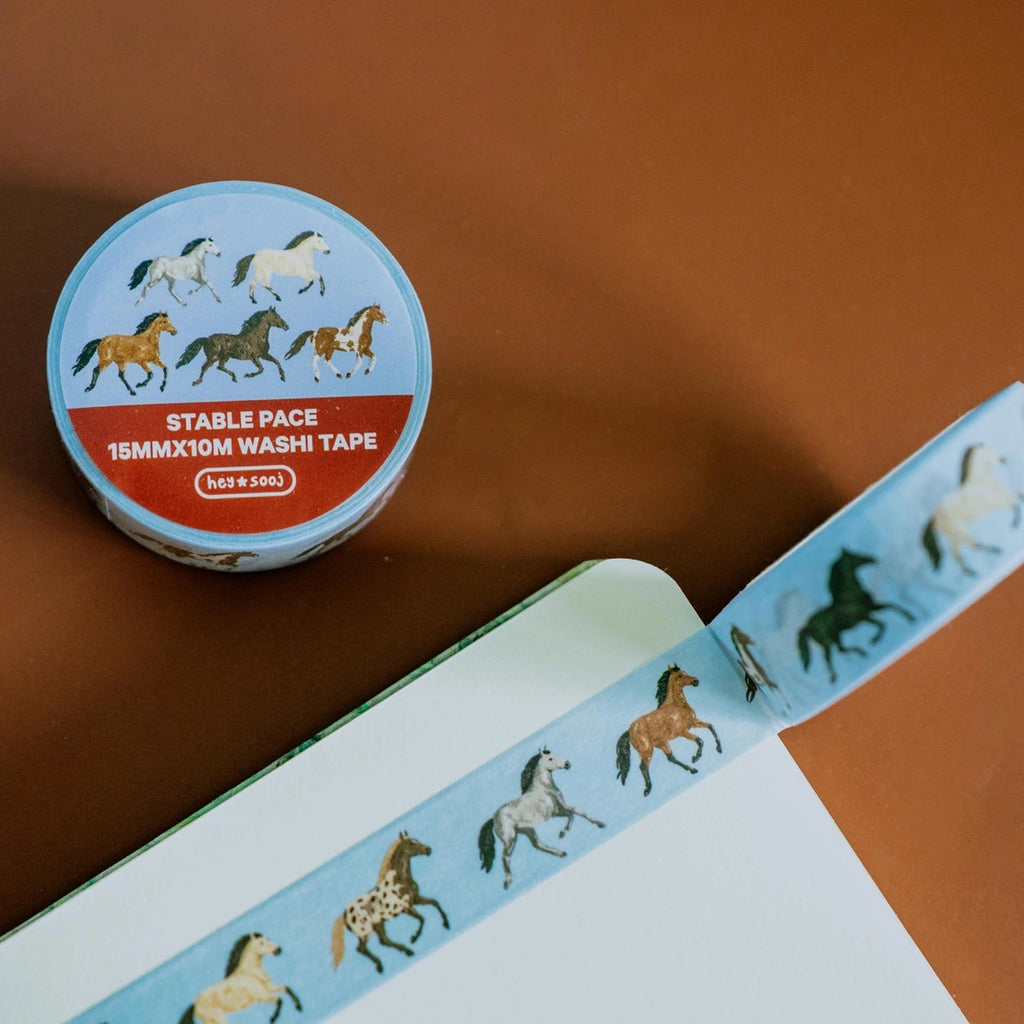 A roll of washi tape with a light blue background and a repeating pattern of horses in various colors, shown partially unrolled next to its circular packaging on a warm brown surface.