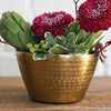 A decorative hammered gold metal bowl filled with a floral arrangement, including two large deep red flowers, green leaves, and small green berries, set on a wooden surface against a white brick wall background.