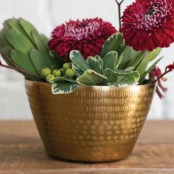 A decorative hammered gold metal bowl filled with a floral arrangement, including two large deep red flowers, green leaves, and small green berries, set on a wooden surface against a white brick wall background.